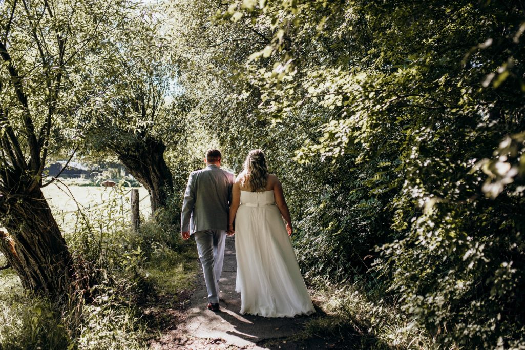 Fotoshooting geplant von Hochzeitsplanerin Nina Hübner Göttingen Brautpaar geht Hand in Hand einen Waldweg entlang, umgeben von üppigem Grün.